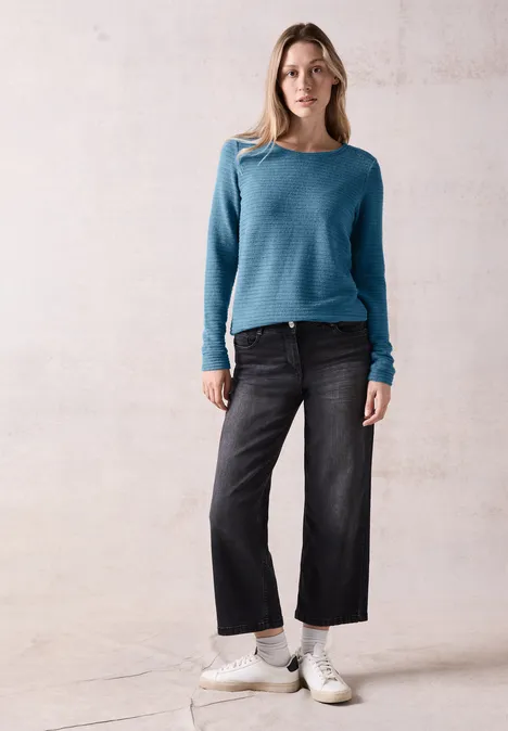 Frau mit blauem Pullover, schwarzer Jeans und weißen Sneakern steht vor hellem Hintergrund.